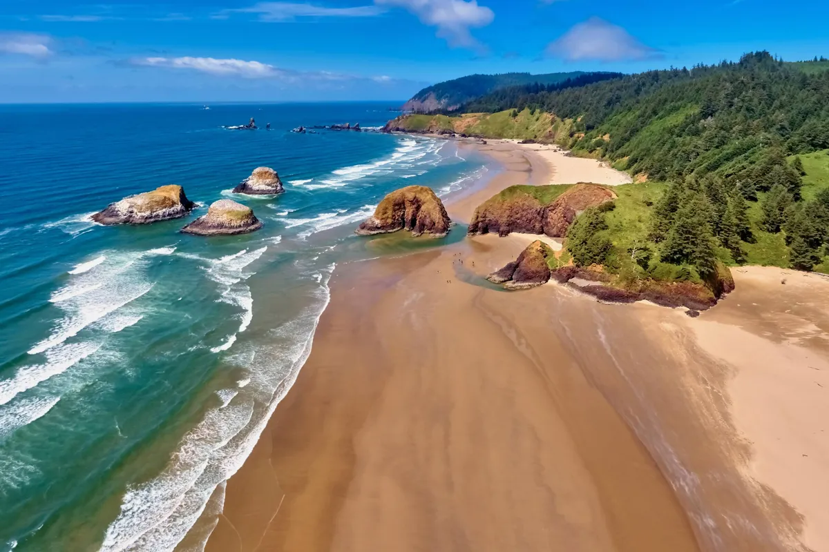 A scenic coastal view with dramatic rock formations, sandy beach, rolling ocean waves, and green hills in the background. The landscape is a quintessential Oregon Coast scene, inviting beachcombing, agate hunting, and exploration.