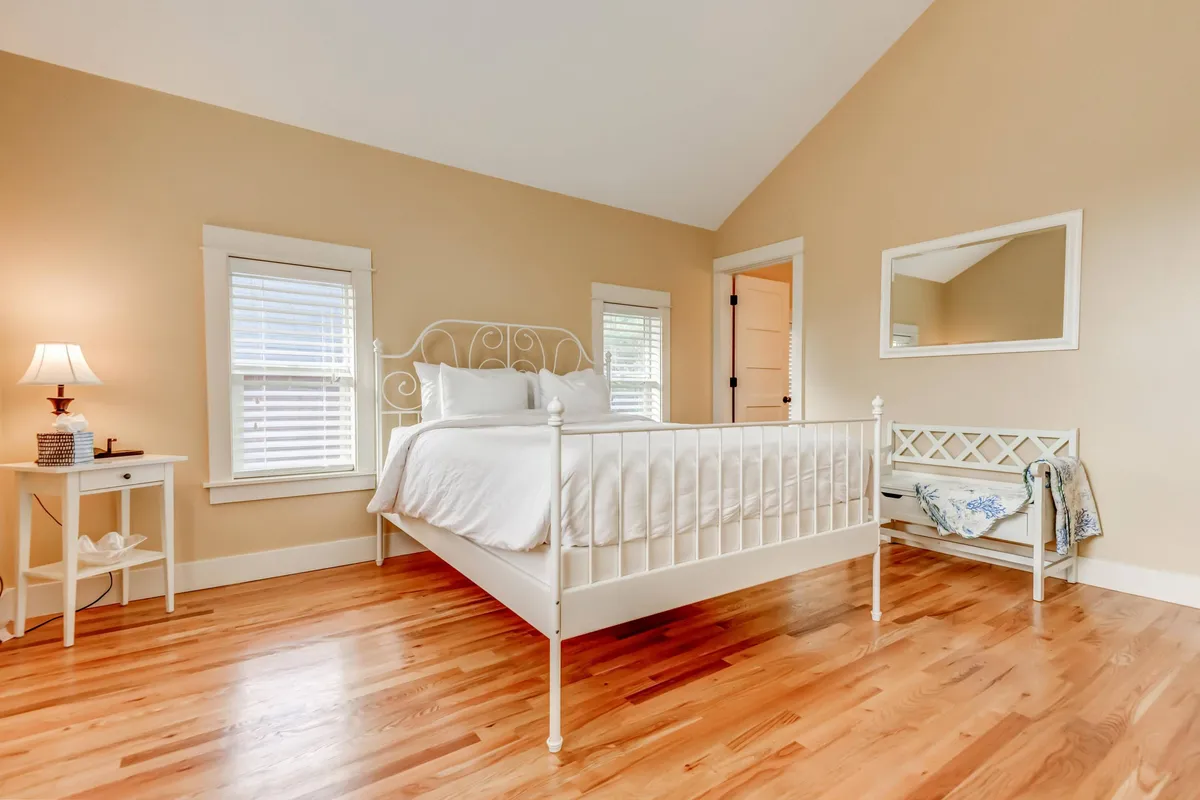 A serene bedroom with white linens, a nightstand and lamp, and a large mirror, all set on wooden flooring. The space invites relaxation and rejuvenation, with soft lighting suggesting a peaceful morning or tranquil night.