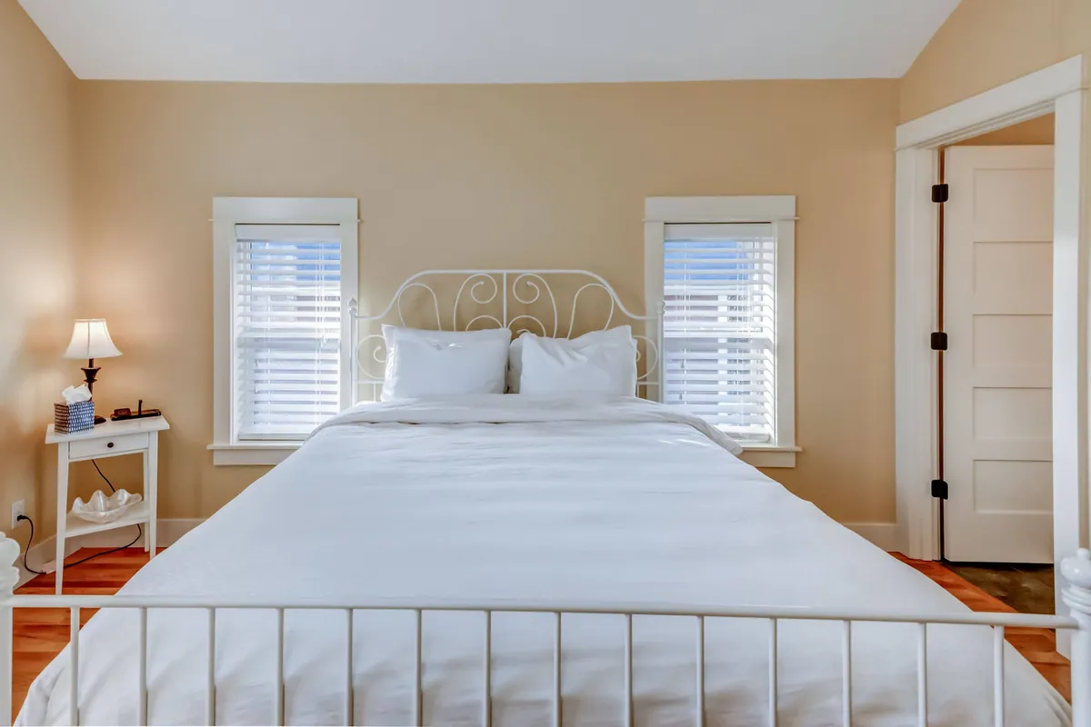 A cozy bedroom with a queen-sized bed dressed in crisp white linens, a nightstand with a warm lamp, and generous windows fitted with blinds. The soft light and neutral palette create an atmosphere of calm, perfect for relaxation after a coastal adventure.