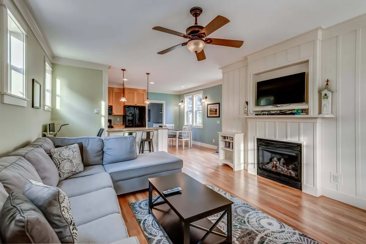 A spacious open-concept living area featuring a comfortable sectional sofa oriented towards a modern fireplace and a large TV. The dining area is seamlessly connected, visible in the background, creating a unified space for relaxation and meals. Natural light streams in, adding warmth to the cozy atmosphere.