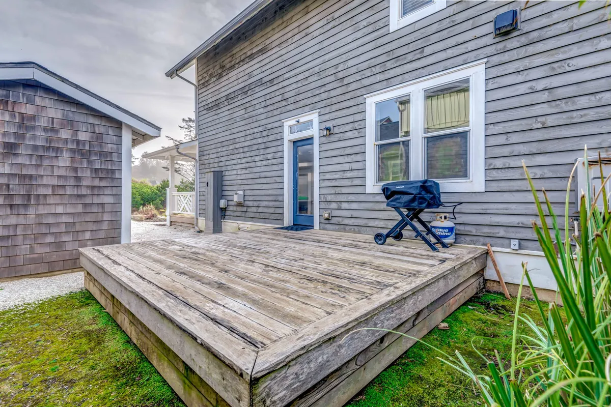 A relaxed wooden deck with a grill and seating area, set against the textured exterior wall of the house. The scene exudes a cozy, welcoming vibe perfect for outdoor gatherings or quiet mornings, with hints of sunset light adding warmth to the atmosphere.
