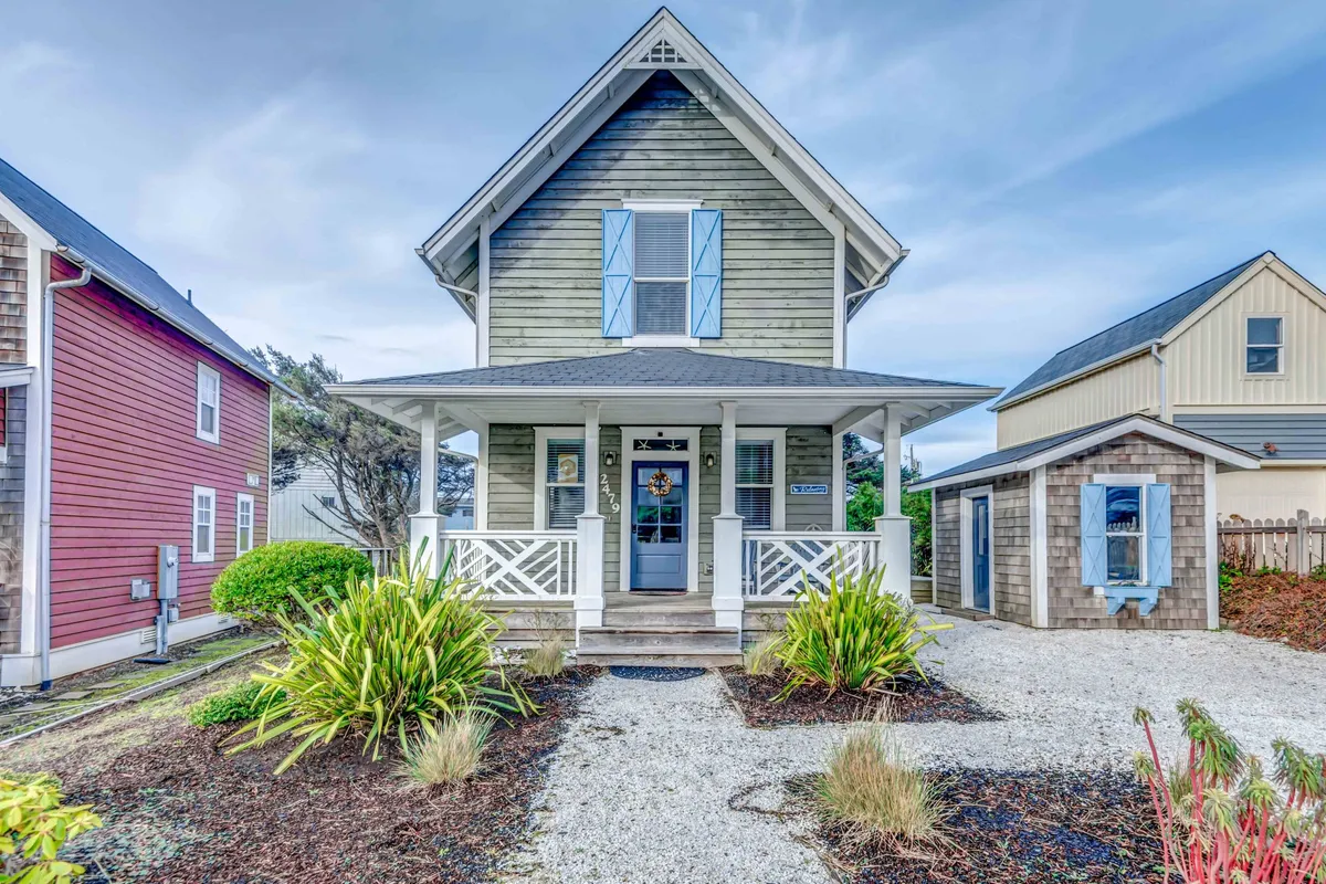 The image showcases the charming exterior of a single-family beach cabin in Lincoln City's upscale Olivia Beach community. The view highlights a welcoming front porch with comfortable seating, surrounded by manicured landscaping and symmetrical architectural details that evoke a sense of tranquility and style. The inviting porch, lush garden, and classic design create an ideal spot for relaxing with a morning coffee or enjoying vibrant sunsets.