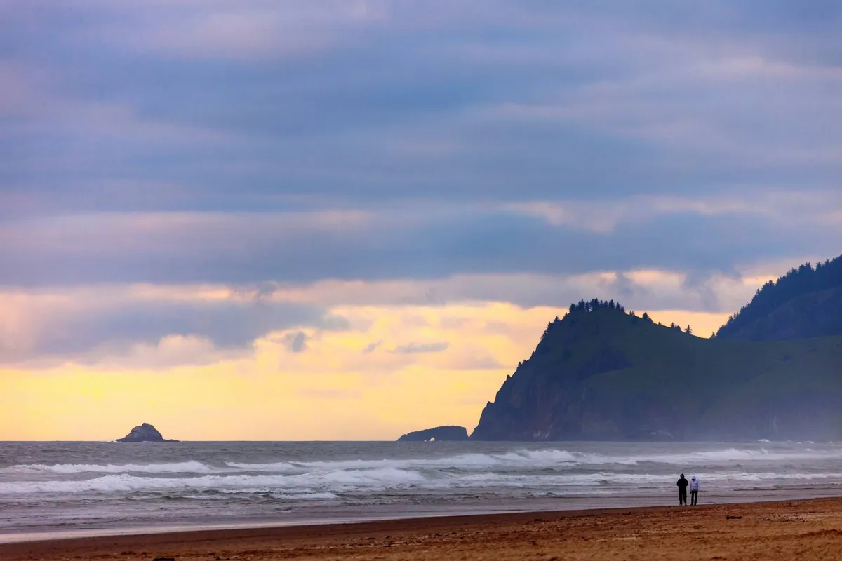 A sweeping view of the ocean with distant cliffs under a colorful sky. In the foreground, a couple walks along the sandy beach, their figures silhouetted by the sunset. The scene is tranquil yet inviting, evoking the spirit of seaside exploration and group outings in Lincoln City.