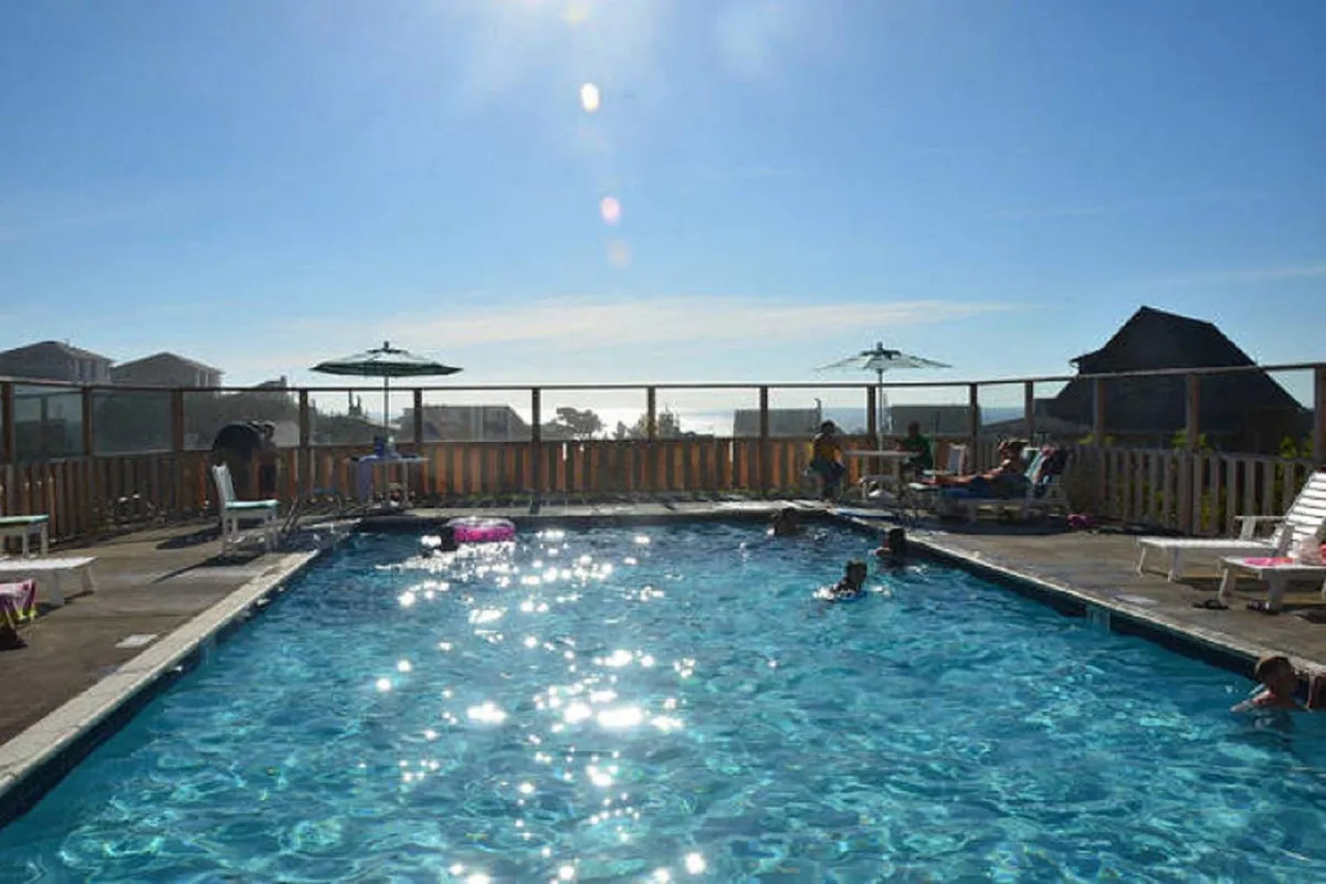 A vibrant outdoor community pool scene with children splashing in the heated saltwater pool, sunbathers relaxing on poolside loungers, and colorful umbrellas offering shade. The pool deck offers panoramic views, and the joyful mood captures summer fun and social interaction.