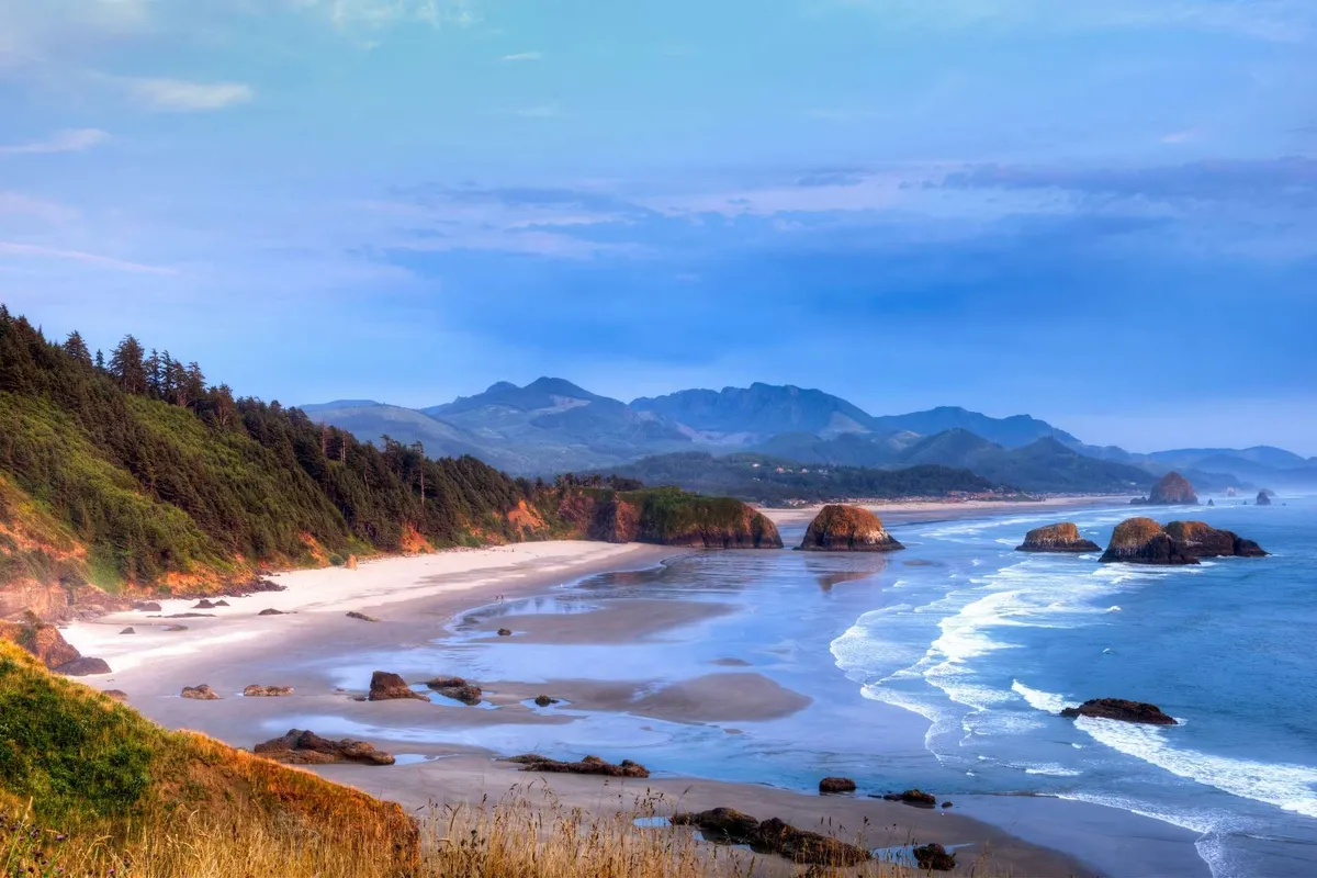 A breathtaking vista of sandy beach, dramatic rock formations in the water, distant mountains, and waves crashing on the shore—an inspiring setting for beachcombing, tide pool exploration, or peaceful strolls.