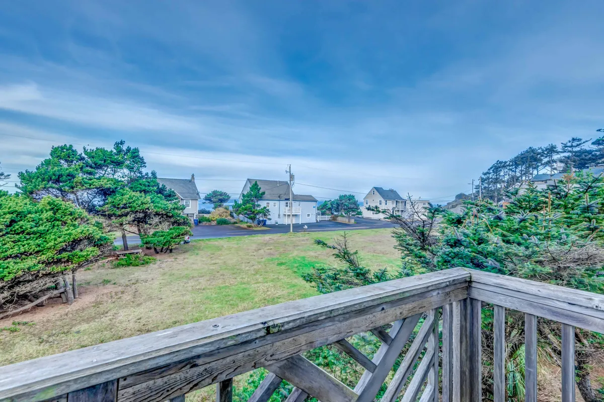 A peaceful view from a wooden balcony overlooking lush greenery, open fields, and distant homes, with a broad sky above. The setting encourages relaxation, casual strolls, and community exploration.