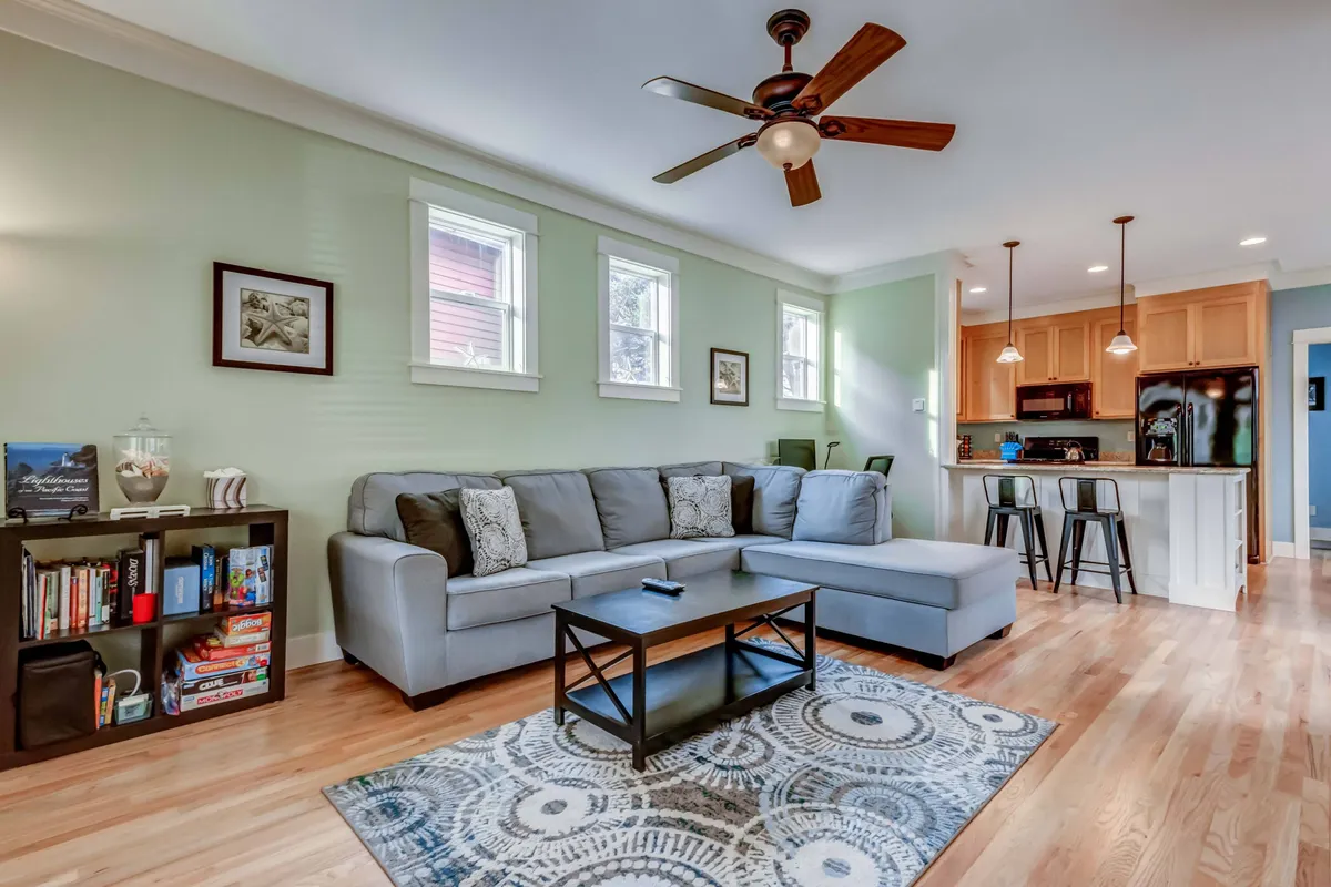 A welcoming living space with a soft sofa, coffee table, bookshelf, and an open view into the kitchen area. The arrangement encourages both relaxation and socializing, with ample light and a homey, well-lived feel.