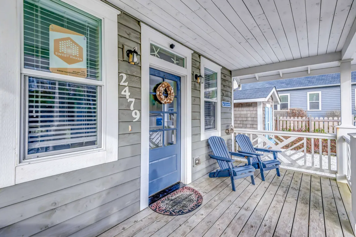 A welcoming exterior shot of the home's front entrance, featuring a charming wreath on the door, a cozy seating area, and visible house number. The porch looks inviting, perfect for relaxing or greeting guests after a day at the beach.